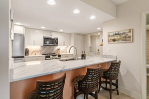 A modern kitchen with a marble countertop, stainless steel appliances, and a bar area with patterned stools. - Tangerine Tides: Resort Pool/Spa, On Beach, Lanai (Fort Myers Beach)