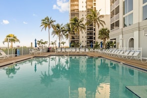 A swimming pool with lounge chairs and palm trees, surrounded by a multi-story building. - Tangerine Tides: Resort Pool/Spa, On Beach, Lanai (Fort Myers Beach)
