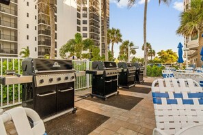 Outdoor barbecue grills, lounge chairs, and a poolside area with palm trees and high-rise buildings in the background. - Tangerine Tides: Resort Pool/Spa, On Beach, Lanai (Fort Myers Beach)
