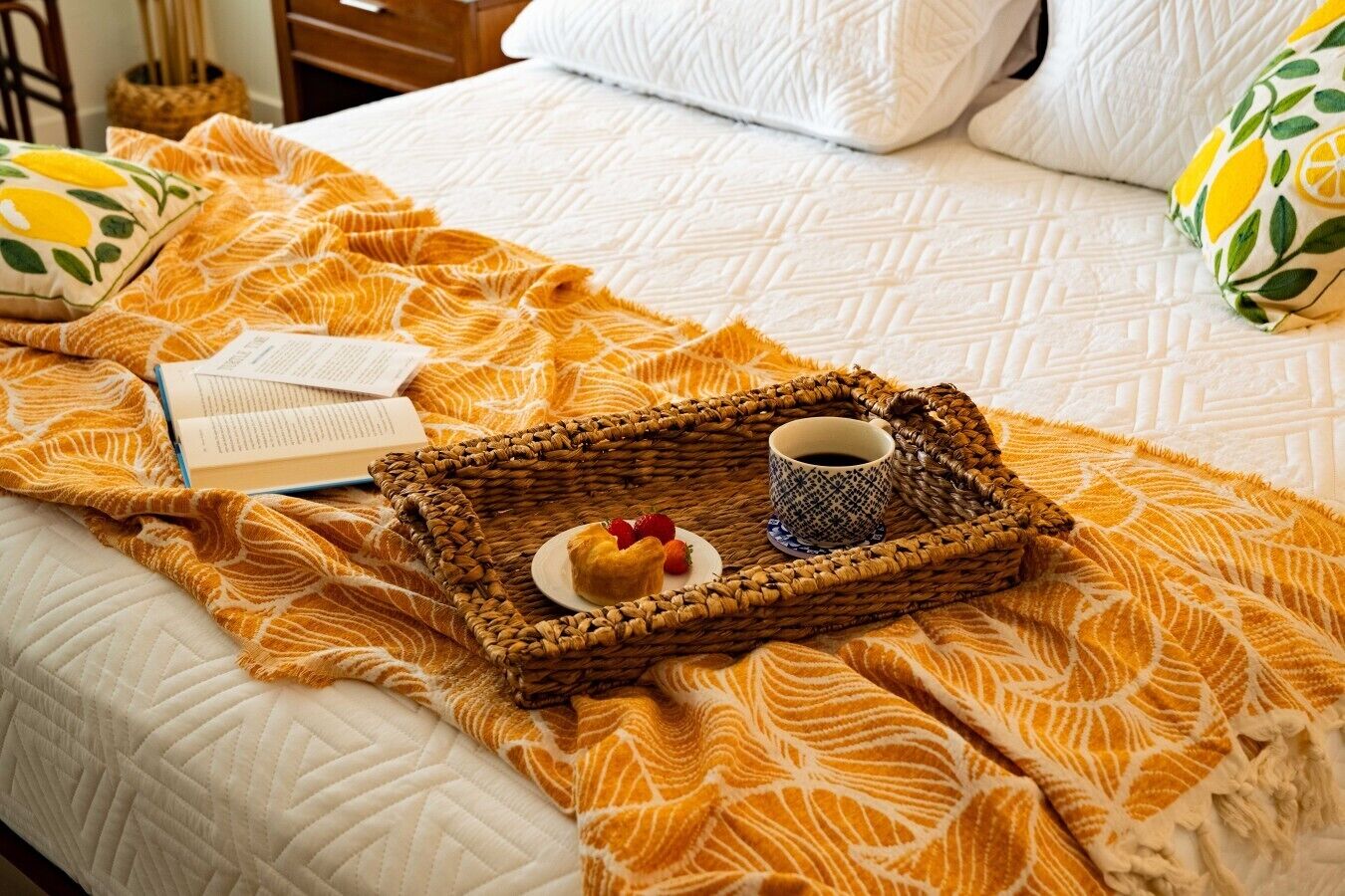 Un lit recouvert d’une couette blanche, d’une couverture à motifs jaunes, une petite table en osier avec une tasse de café, une pâtisserie et un livre.