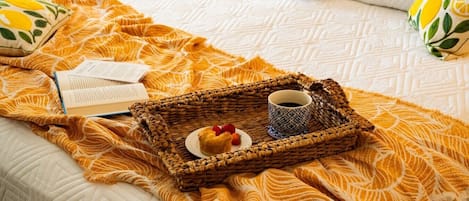 A bed with a white quilt, a yellow patterned blanket, a wicker tray with a cup of coffee, a pastry, and a book.