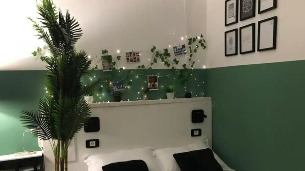 Un dormitorio con una cama, almohadas negras, una planta en maceta, luces fijadas a la pared y cuadros enmarcados.