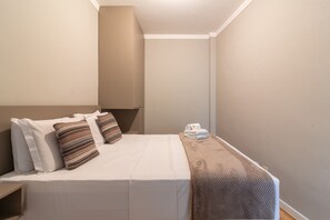 Ein kleines Zimmer mit einem Einzelbett, einer schlichten weißen Bettdecke, zwei Kissen, einem gefalteten Handtuch und einer Decke auf dem Bett.