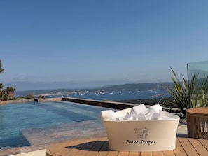 An infinity pool with a view of the sea and distant hills, a white bucket labeled 'Saint Tropez,' and a glass table with plants. - Villa Domaine du Col: Pool, Jacuzzi, Exceptional View, 5 Minutes from the Beaches (Grimaud)
