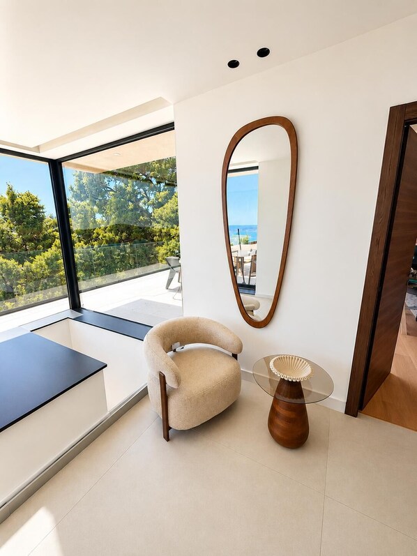 A modern interior with a large window, a round mirror, a beige armchair, and a glass table.