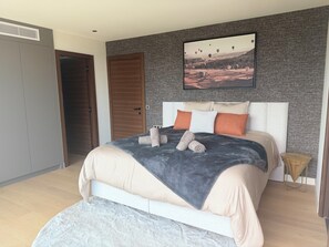 Un dormitorio con cama, almohadas, una colcha, cabecera, un cuadro en la pared y una puerta de madera.