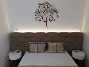 Ein Schlafzimmer mit Kopfteil, zwei Nachttischen, einem Bett mit kariertem Kissen und einer Baum-Wanddekoration.