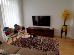 Ein Wohnzimmer mit einer Couch, zwei Sesseln, einem Fernseher, einer Holzkommode, einem gemusterten Teppich und einer Topfpflanze.