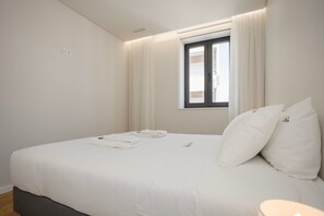 Una cama bien hecha con sábanas blancas, una ventana con cortinas y una mesita de noche con lámpara.