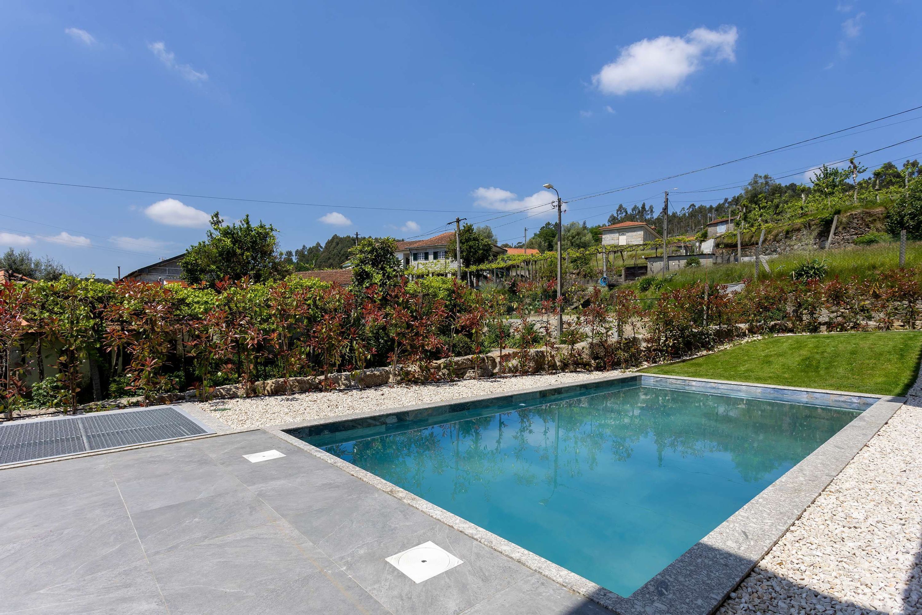 Una piscina con bordeado de baldosas, rodeada por un jardín ajardinado con diversas plantas y una colina al fondo.