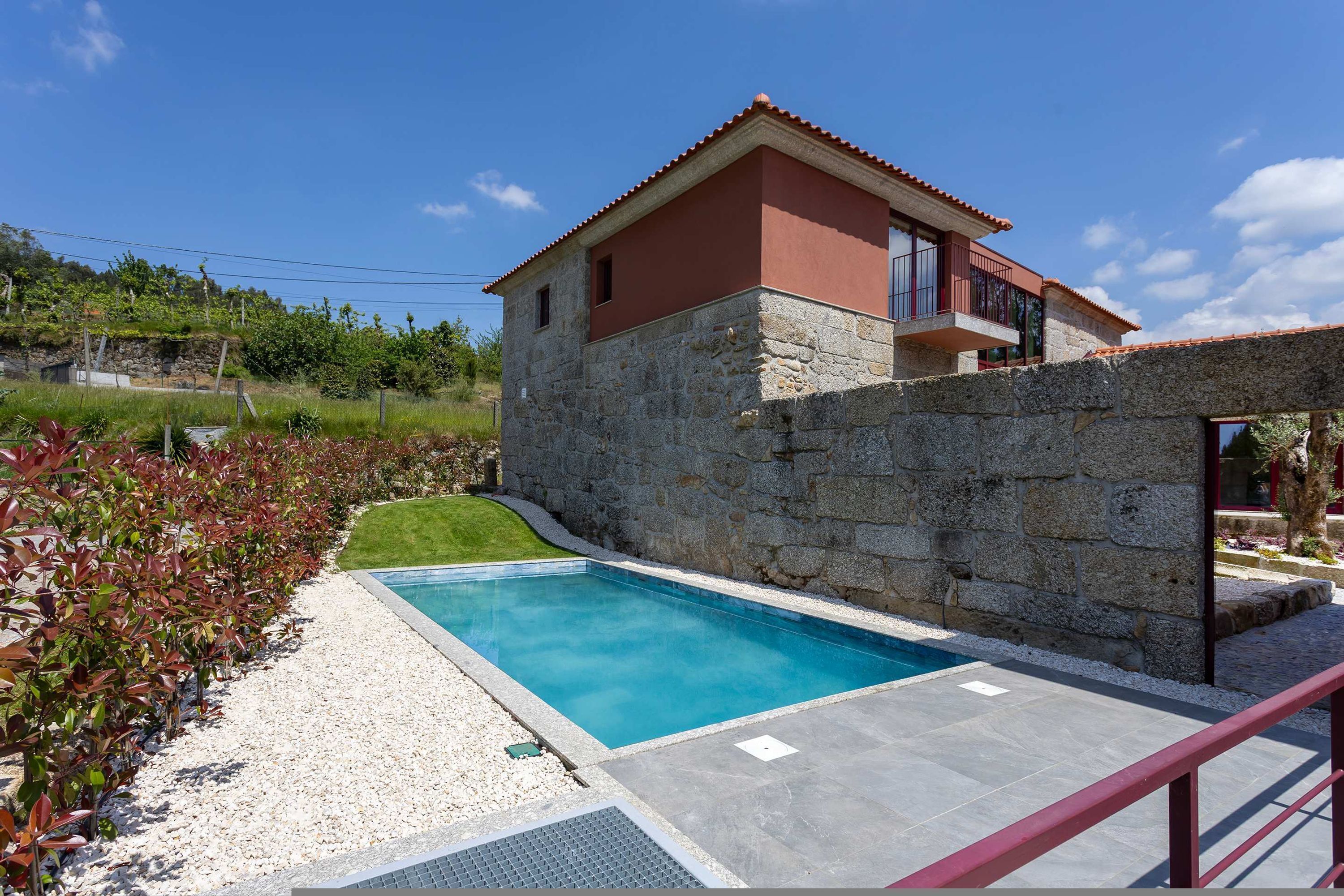 A two-story stone house with a swimming pool, surrounded by a garden and a stone wall.