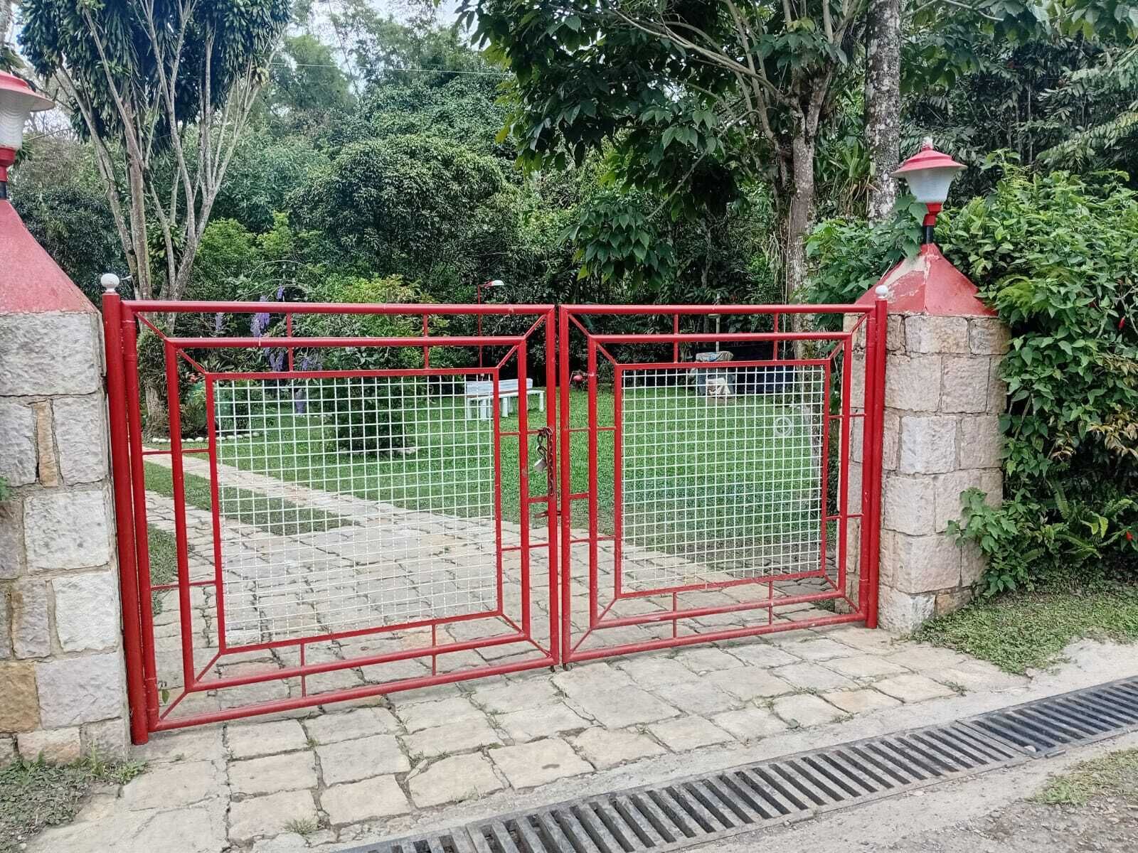 A red metal gate with a stone wall, a grassy area beyond, and a tree in the background.