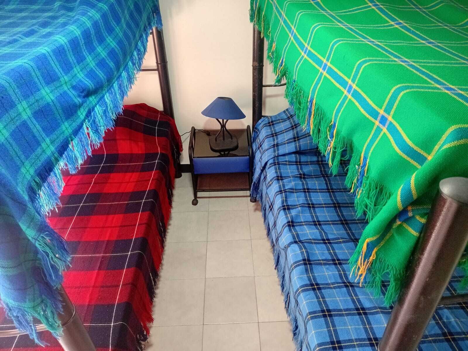 A dormitory room with bunk beds, a small table with a lamp, and a chair.