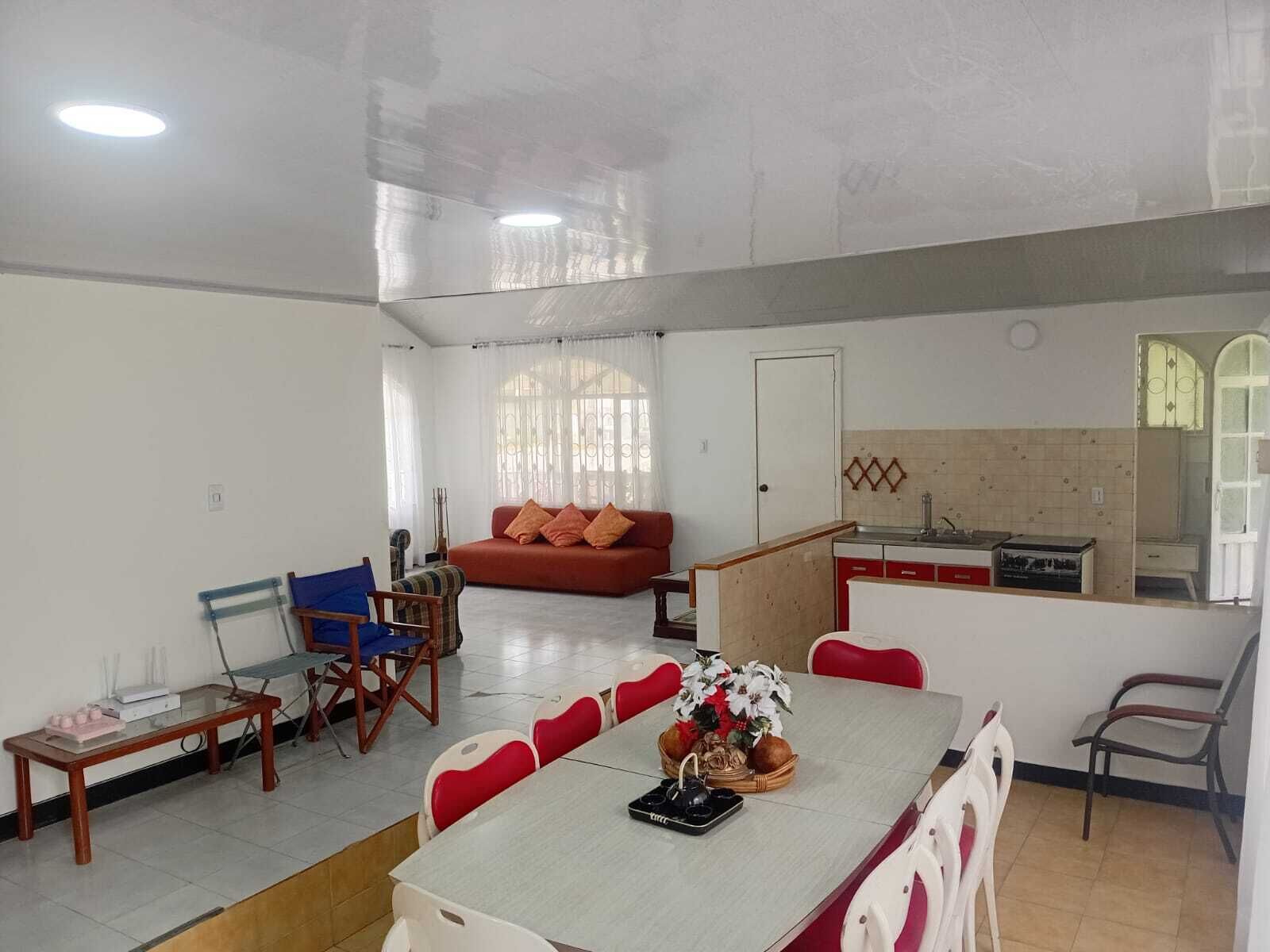 A spacious living area with a dining table, red and white chairs, a sofa, and a kitchen area with a tiled backsplash.