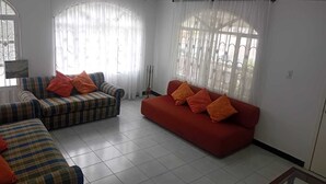 A living room with two sofas, one striped and one solid red, both with orange cushions. There are large windows with sheer curtains, and a tiled floor.