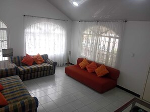 A living room with two sofas, one striped and one solid red, both with orange cushions. There are large windows with sheer curtains, and a tiled floor.