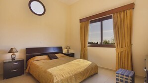 A bedroom with a bed, two bedside tables, a round mirror, a window with curtains, and a small bench. - Villa Petra Sun-Coral Bay (PAFOS)