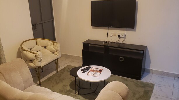 A living area with a sofa, a chair, a small table with a remote control, and a TV mounted on the wall. - Cozy 1-bedroom apartment with AC in pleasant Machakos (Machakos)