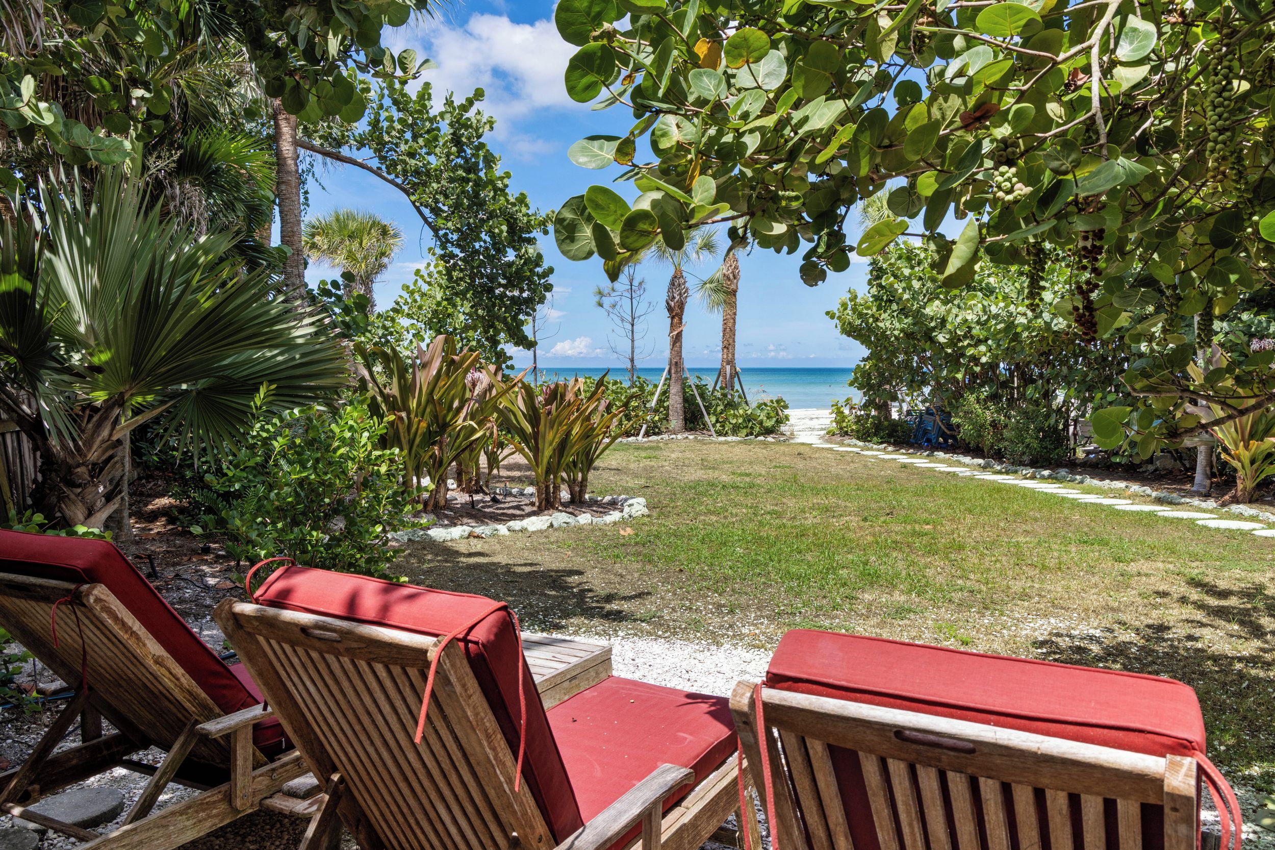 Un jardín tropical con vista al océano, sillones de madera y un camino que lleva a la playa.