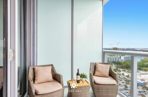 Un balcón con dos sillones de mimbre, una bandeja con una botella y vasos, y vistas de un paisaje urbano con edificios y cielo despejado.