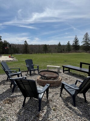 A fire pit surrounded by outdoor seating, a basketball hoop, and a grassy field with trees in the background. - Family friendly 3-bedroom cabin in peaceful Vanderbilt. Private. Large groups. (Vanderbilt)
