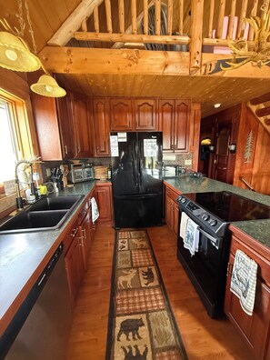 A wooden cabin interior with a black refrigerator, wooden cabinets, a stove, and a rug with animal prints. - Family friendly 3-bedroom cabin in peaceful Vanderbilt. Private. Large groups. (Vanderbilt)