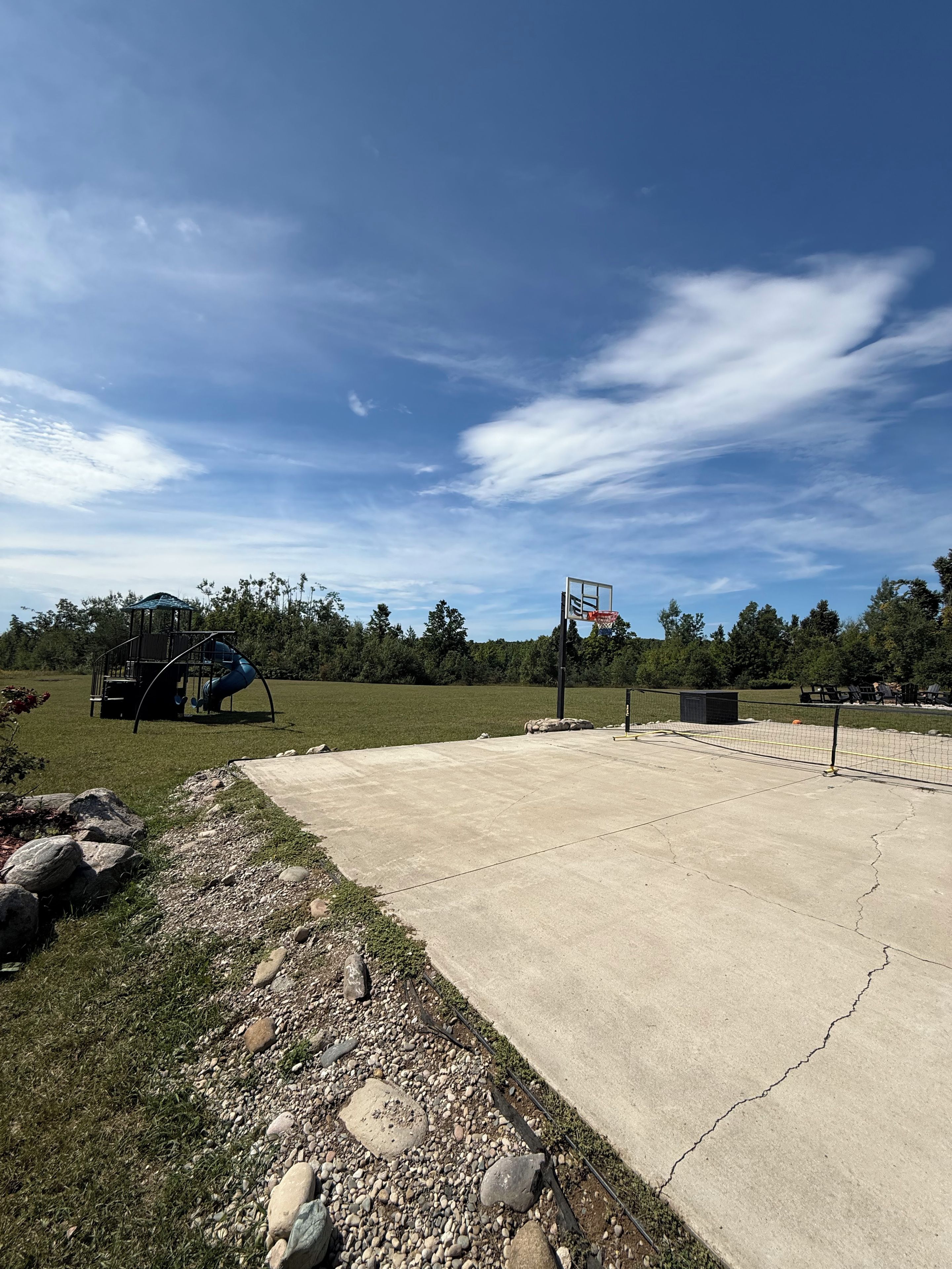 Una cancha de baloncesto de concreto con un aro, rodeada de césped y árboles.