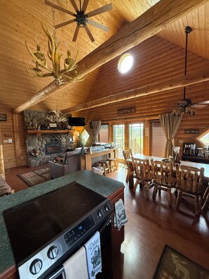 A cabin interior with a stone fireplace, wooden dining table, and a kitchen area with a stove. - Family friendly 3-bedroom cabin in peaceful Vanderbilt. Private. Large groups. (Vanderbilt)