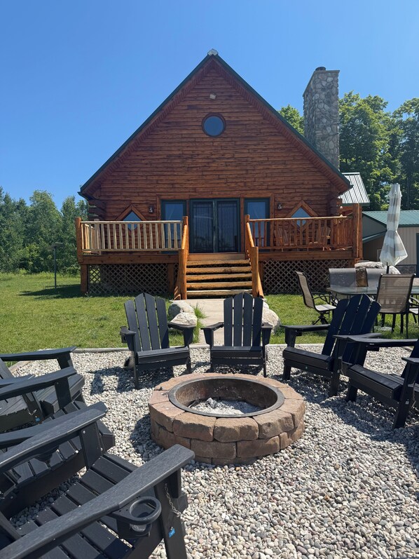 A wooden cabin with a stone chimney, a deck with chairs, and a fire pit surrounded by Adirondack chairs.