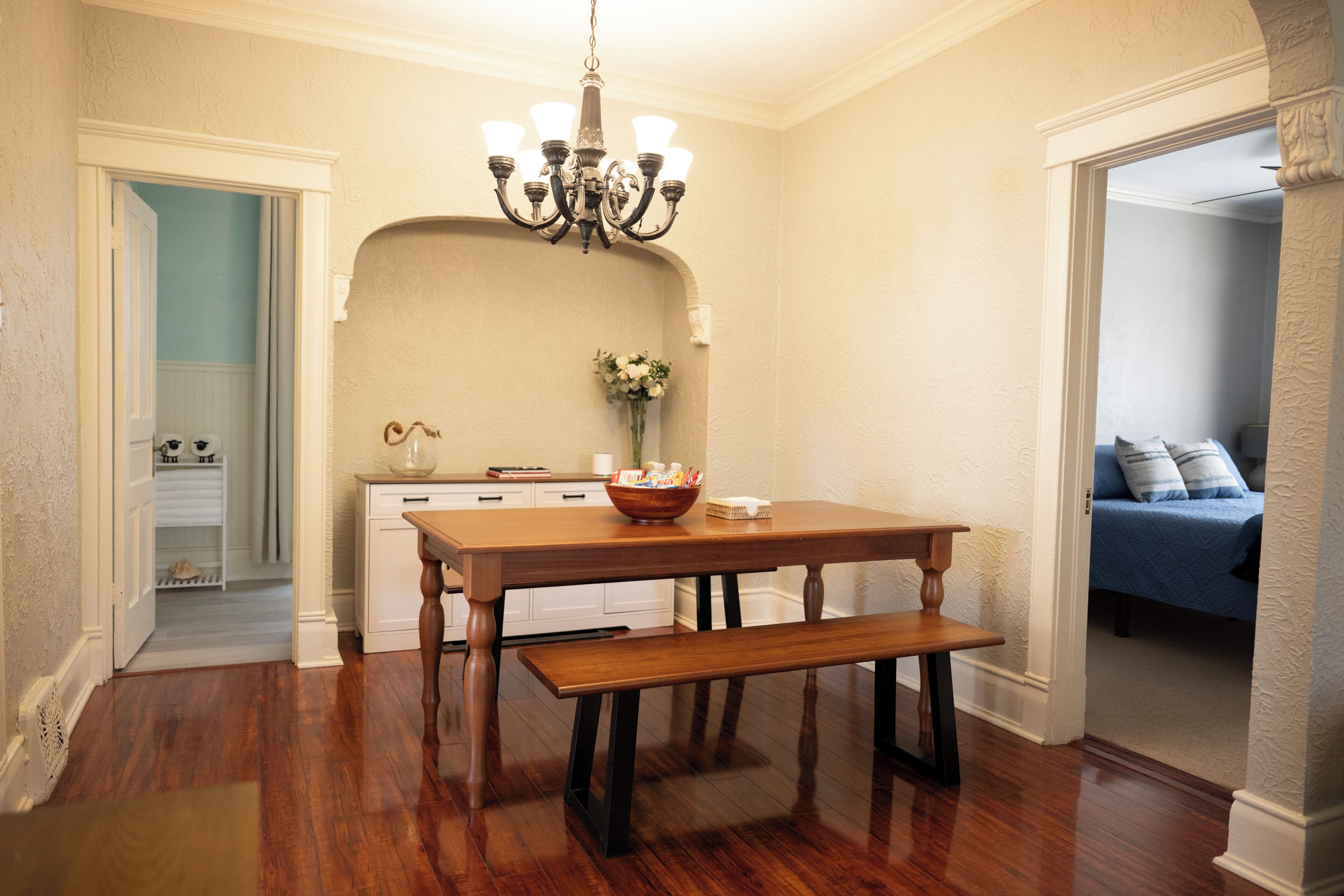 Comedor con mesa y banco de madera, una lámpara de araña y un florero con flores.