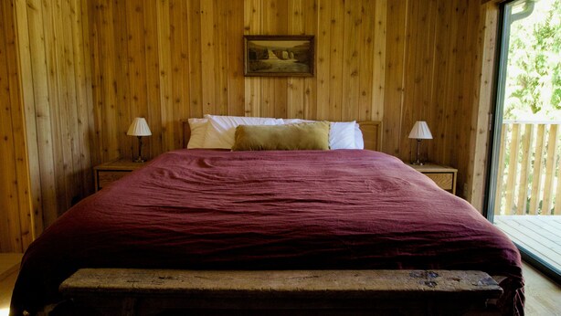 Quarto com painéis de madeira, uma cama, duas mesas de cabeceira, um banco e uma janela.