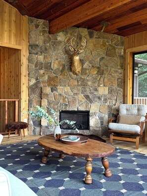 A stone fireplace with antlers mounted above it, a wooden ceiling, a patterned rug, a round wooden table, and a cushioned chair. - Lakeview 4BR home, Wellness Spa, Cedar Forest, 7 Min to Town- kumo cabin! (Sagle)