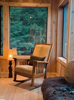 Ein raumförmiger Innenraum mit Holzbalken, einem großen Fenster, einem Holzstuhl, einem kleinen Tisch und einer Lampe.