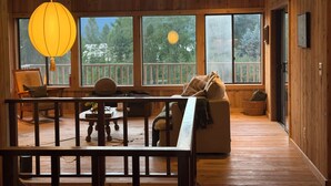 Interior de uma cabana de madeira com uma janela grande, um candeeiro suspenso, um sofá e uma mesa de jantar.