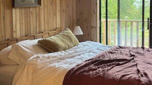 Quarto com parede de painéis de madeira, cama com roupa de cama branca e manta marrom, mesa de cabeceira, abajur e janela com vista para árvores.
