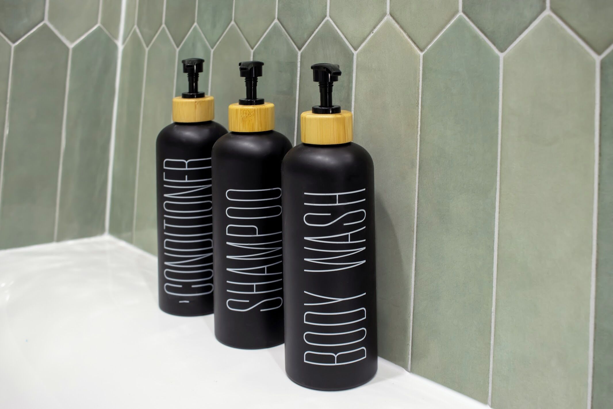 Tres botellas negras con tapas amarillas etiquetadas 'BODY WASH' sobre una superficie blanca, frente a una pared de azulejos verdes.