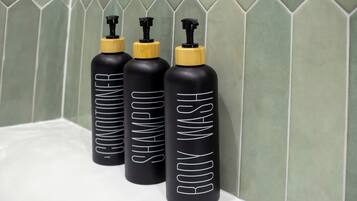Tres botellas negras con tapas amarillas etiquetadas 'BODY WASH' sobre una superficie blanca, frente a una pared de azulejos verdes.