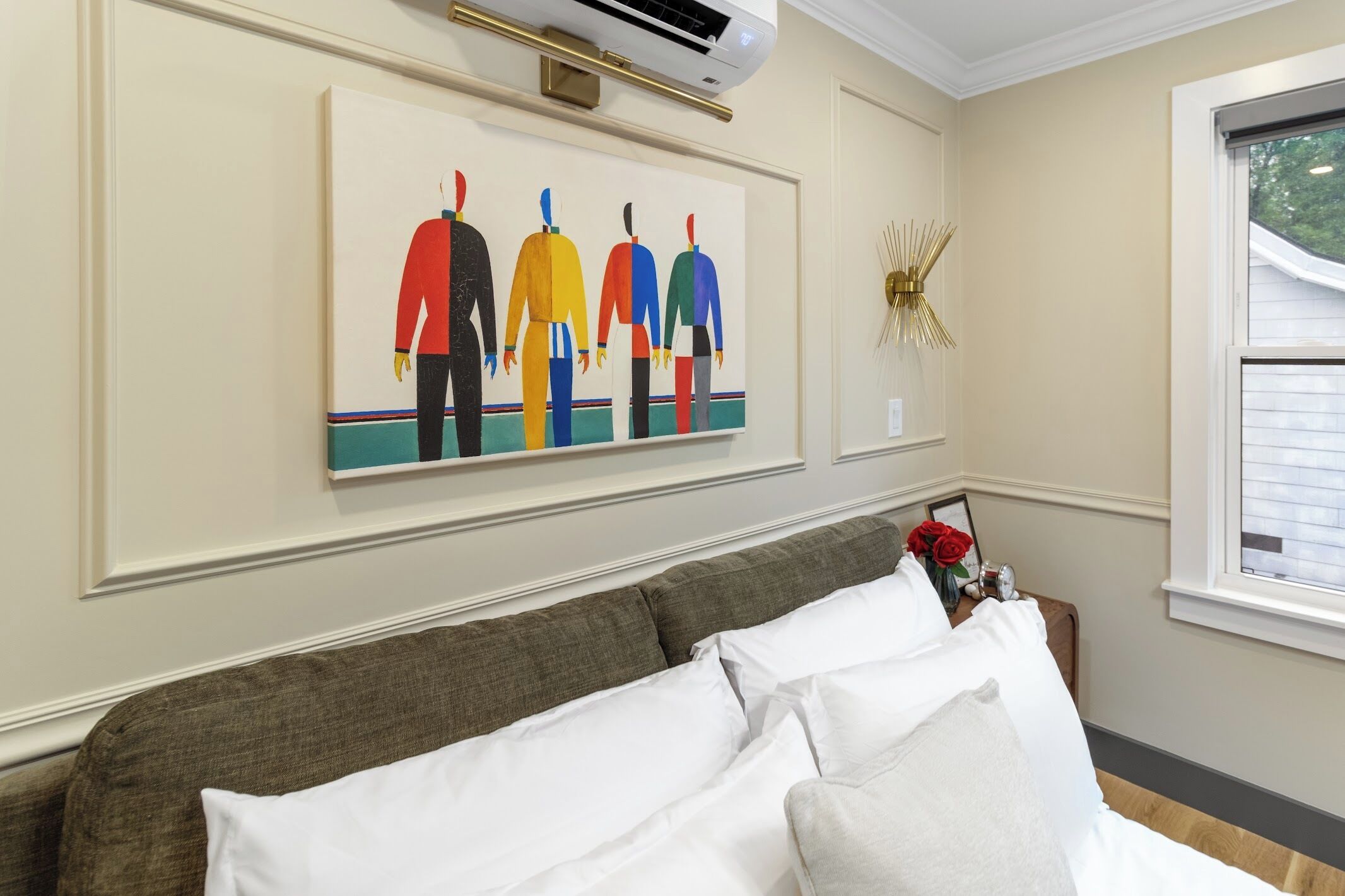 A room with a bed, a wall-mounted air conditioner, a framed artwork, and a window with blinds.
