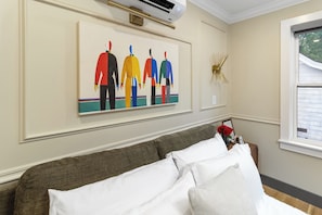 A room with a bed, a wall-mounted air conditioner, a framed artwork, and a window with blinds.