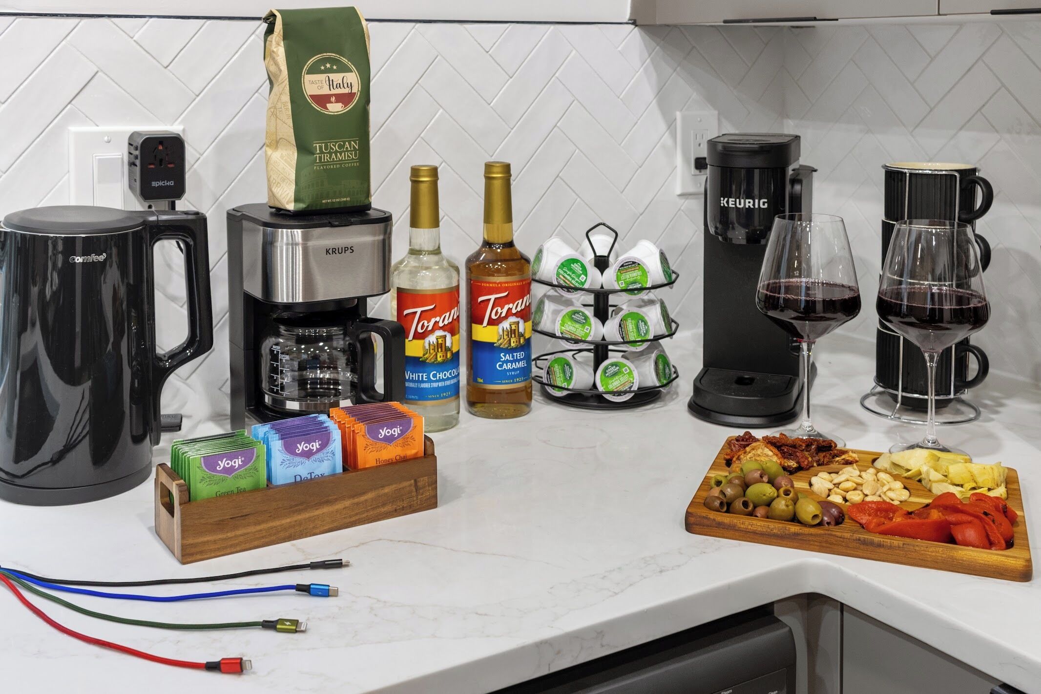 A kitchen counter with a coffee maker, a kettle, a coffee machine, two bottles of Torani syrup, a pack of Yogi tea, a wooden board with snacks, and a fruit platter.
