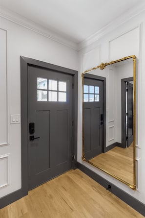 A hallway with a gray door, a large mirror with a gold frame, and a wooden floor.