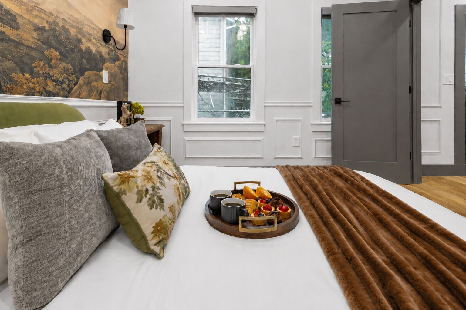 A bedroom with a bed, pillows, a tray of fruits and coffee, a window, and a door.