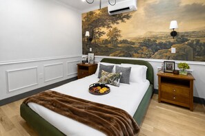 A hotel room with a large bed, a painting on the wall, a nightstand with a lamp, and a tray of fruit on the bed. - Cambridge Designer Home / Harvard, MIT, Boston (Cambridge)