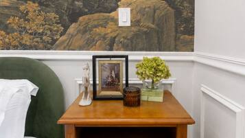 A room with a green sofa, a wooden nightstand with drawers, a lamp, and a landscape mural on the wall.
