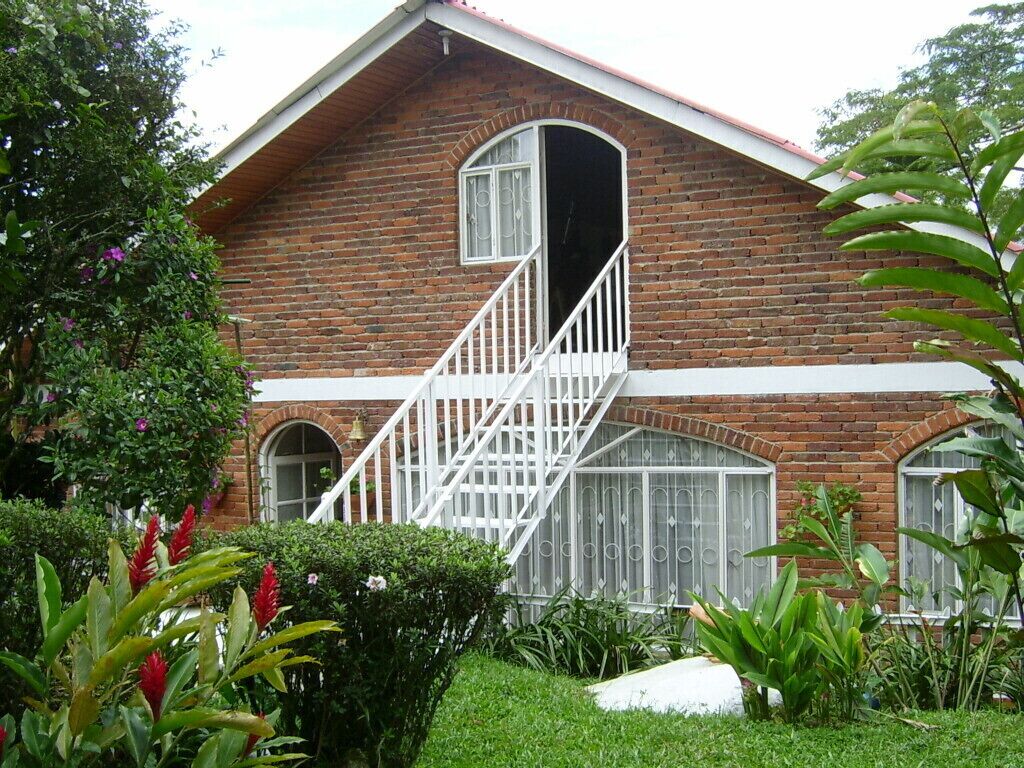 A brick house with a white staircase leading to a balcony, surrounded by lush greenery.