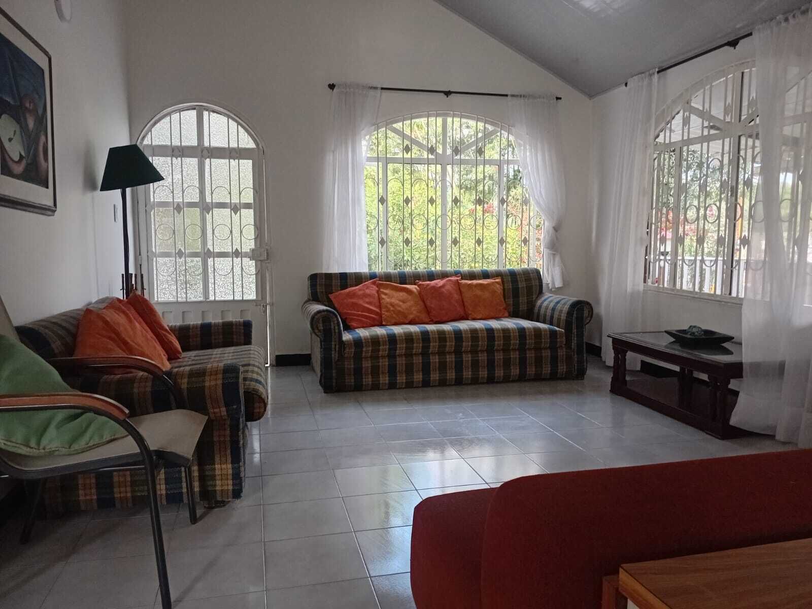 Ein Wohnzimmer mit zwei Sofas, einem Sessel, einem Tisch und großen Fenstern mit Vorhängen.