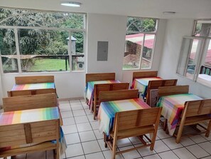 A classroom with wooden benches and tables, large windows, and a view of greenery outside.
