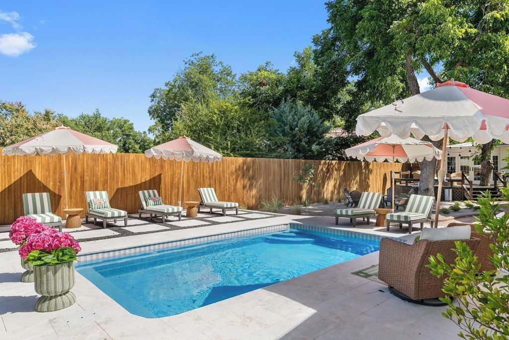A swimming pool with lounge chairs and umbrellas, surrounded by a wooden fence and trees.