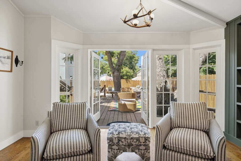 A room with two striped armchairs, a patterned ottoman, a chandelier, and a view of an outdoor deck with furniture and a tree.