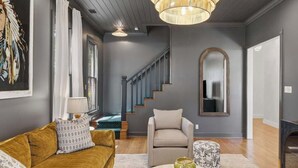 A living room with a sofa, armchair, and ottoman, a staircase, and a large mirror. - Painted Lady - Pool, Hot Tub & Walk to Main (Fredericksburg)